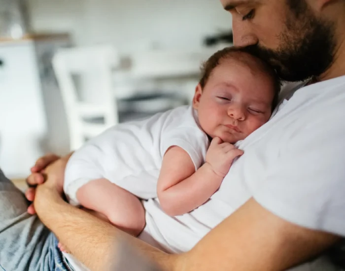 A father kissing his baby on his head while the baby is sleeping on his father’s chest